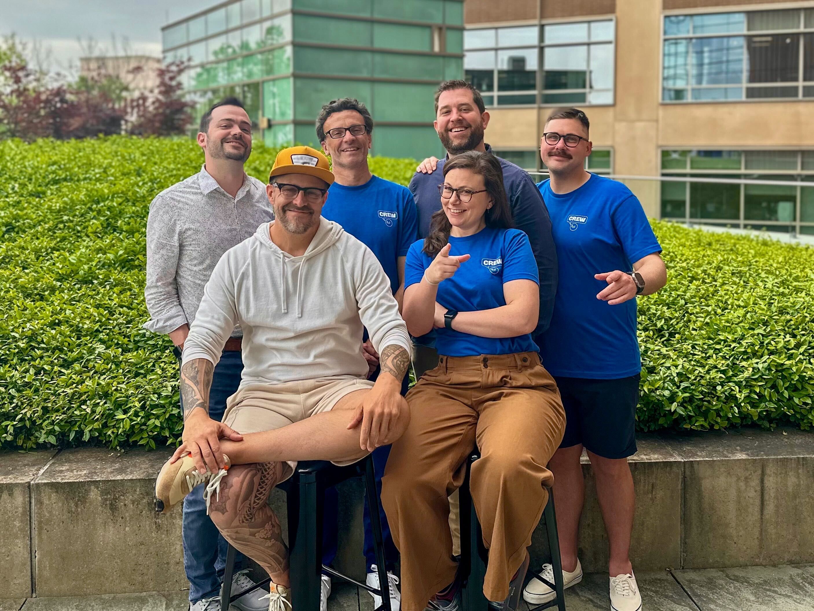 Group of six smiling people posing outdoors; three wear matching blue CREM shirts, one person sits on a stool in front.